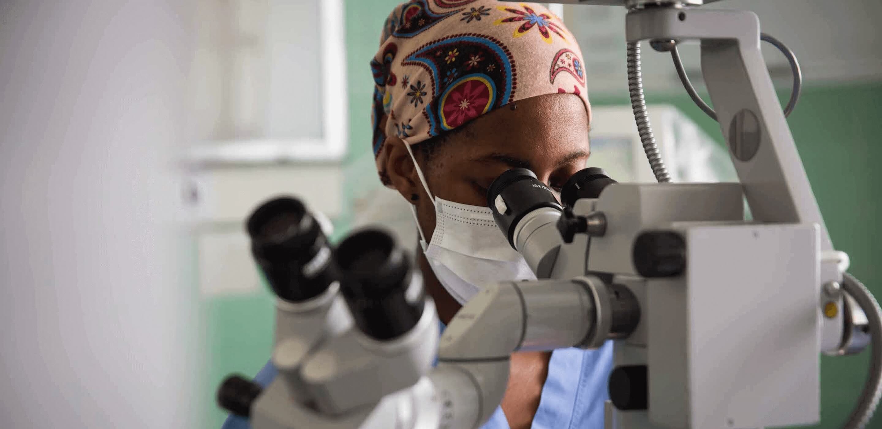 Cancer researcher in scrubs and a mask looking through a microscope.